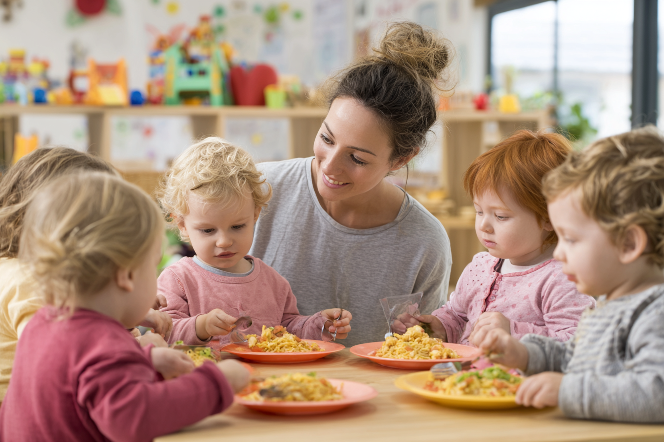 Eine Erzieherin sitzt mit einer Gruppe kleiner Kinder am Tisch und begleitet sie liebevoll beim gemeinsamen Mittagessen in der Kita
